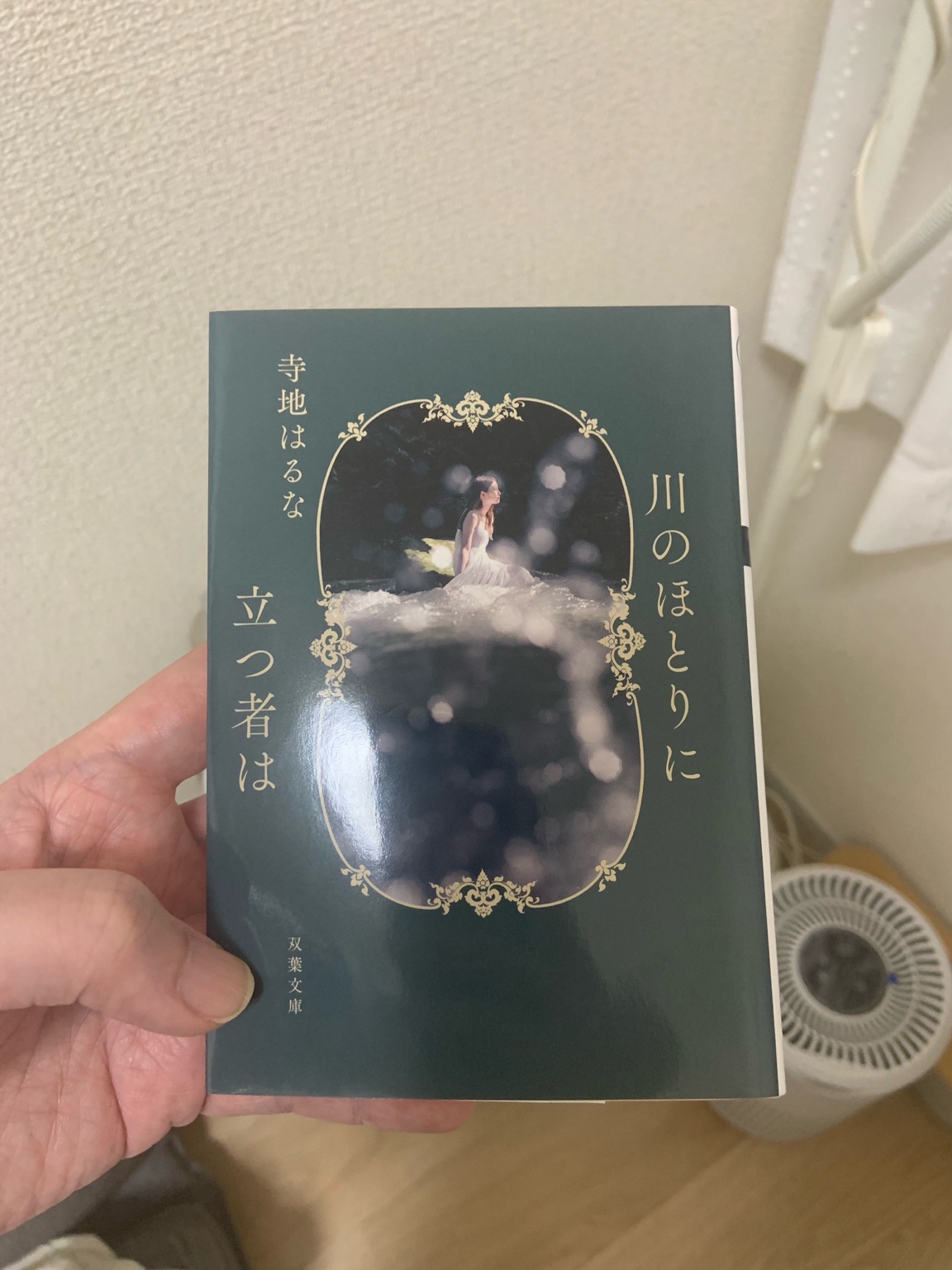 手に持った文庫本『川のほとりに立つ者は』。濃い緑色を背景にしたカバーが印象的