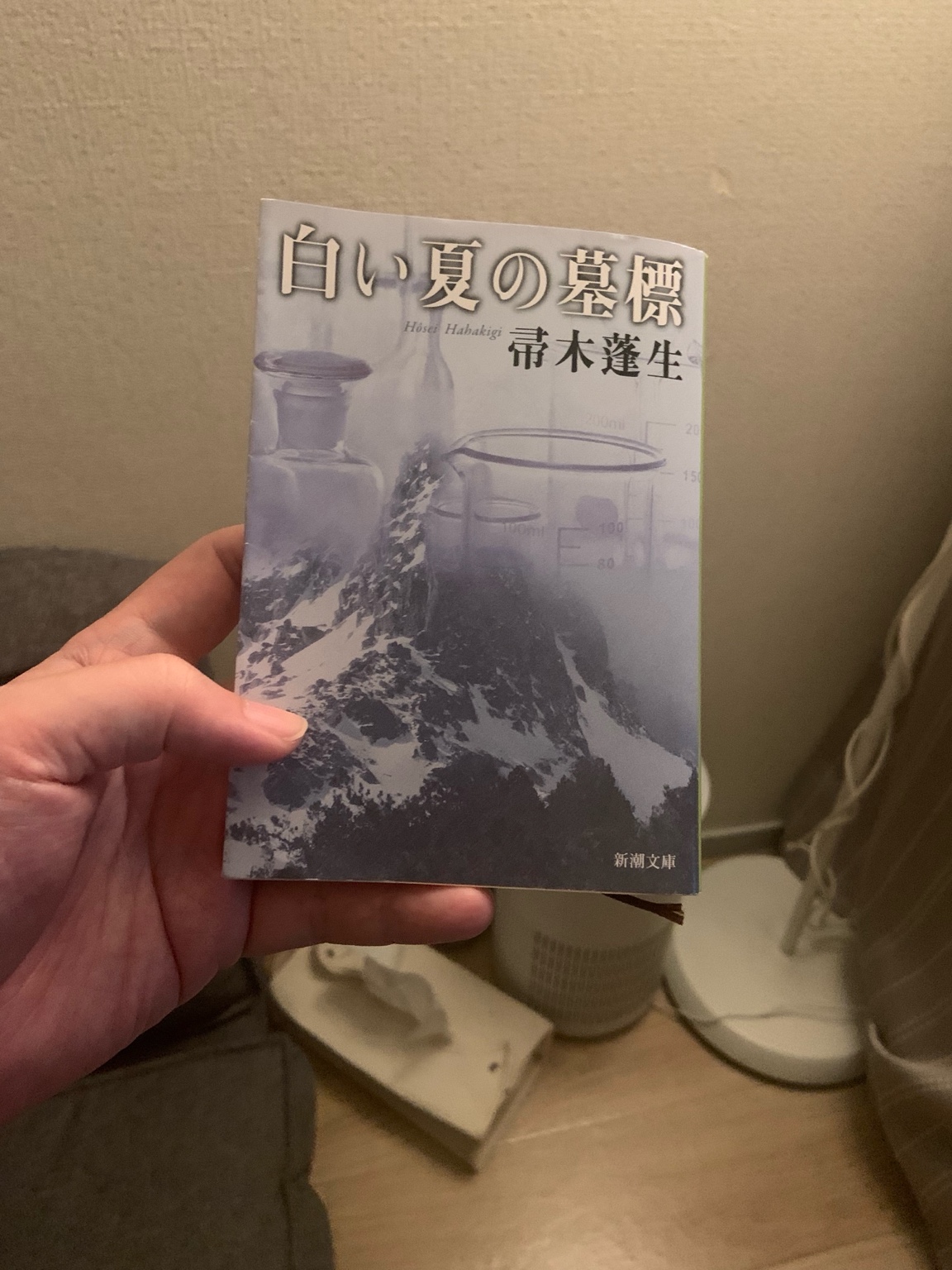 手に持った文庫本『白い夏の墓標』。雪山と理科実験器具が重なった印象的なカバーが特徴。